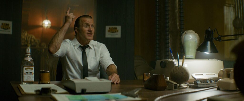 Scott Caan sentado en una oficina con una botella sobre la mesa