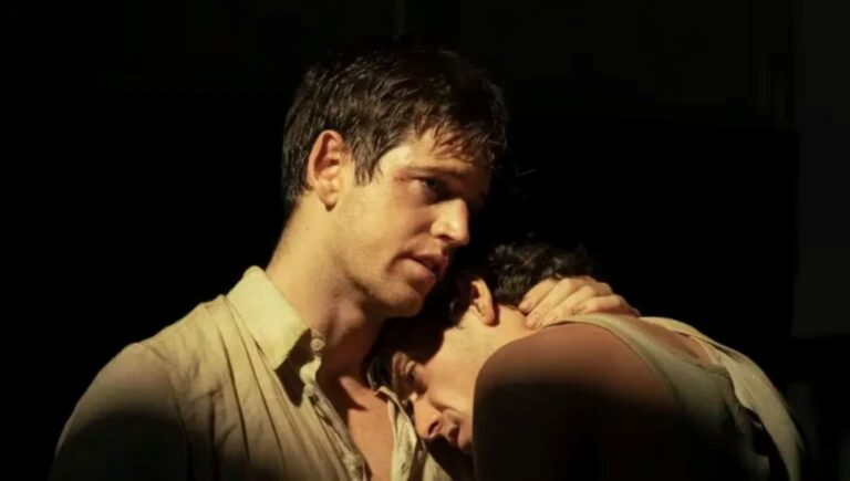 Dos hombres se abrazan cariñosamente en un plano medio con iluminación dramática y fondo oscuro.