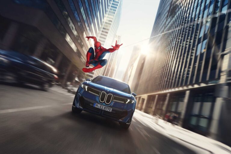 Spider-Man salta sobre un BMW iX3 azul que avanza por una avenida entre rascacielos