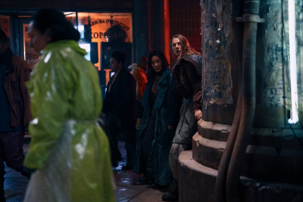 Michelle Yeoh y Hunter Schafer se asoman tras una columna en una calle nocturna y lluviosa en Blade Runner 2099