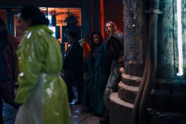 Michelle Yeoh y Hunter Schafer se asoman tras una columna en una calle nocturna y lluviosa en Blade Runner 2099