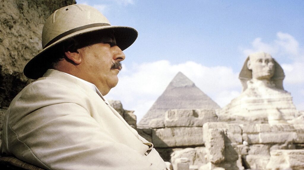 Peter Ustinov como Hércules Poirot, con salacot y traje blanco, ante la Esfinge y una pirámide de Guiza en Muerte en el Nilo (1978)