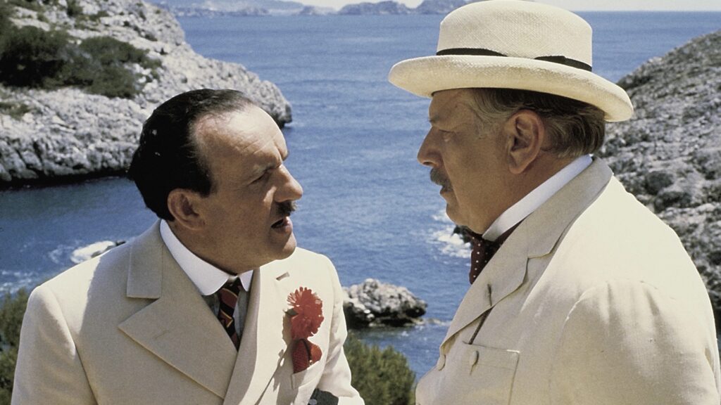 Peter Ustinov como Poirot, con sombrero de paja, conversa con otro personaje vestido de blanco ante una cala en Muerte bajo el sol (1982)