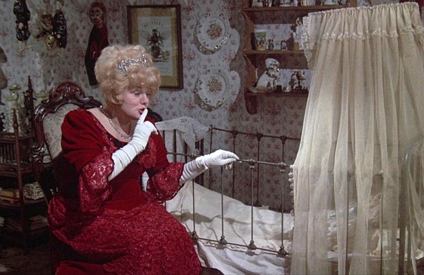 Shelley Winters vestida de gala pide silencio junto a una cuna en una habitación.