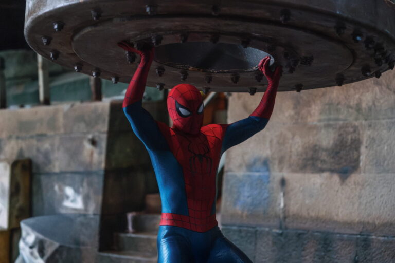 Spider-Man sostiene con los brazos en alto una enorme compuerta metálica entre muros de piedra