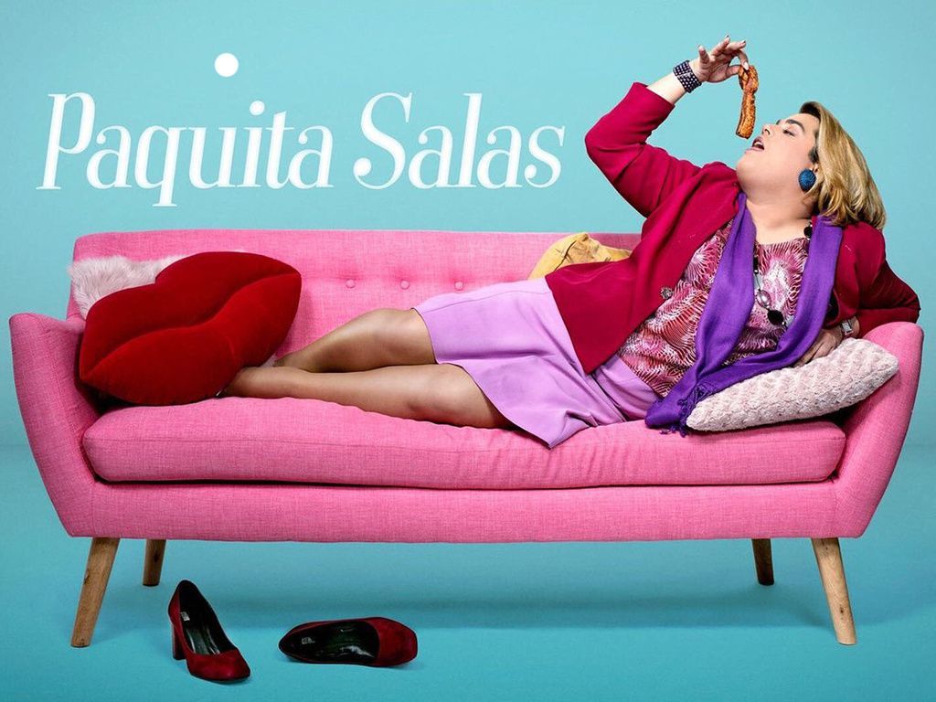 Brays Efe como Paquita Salas tumbada en un sofá rosa comiendo un snack